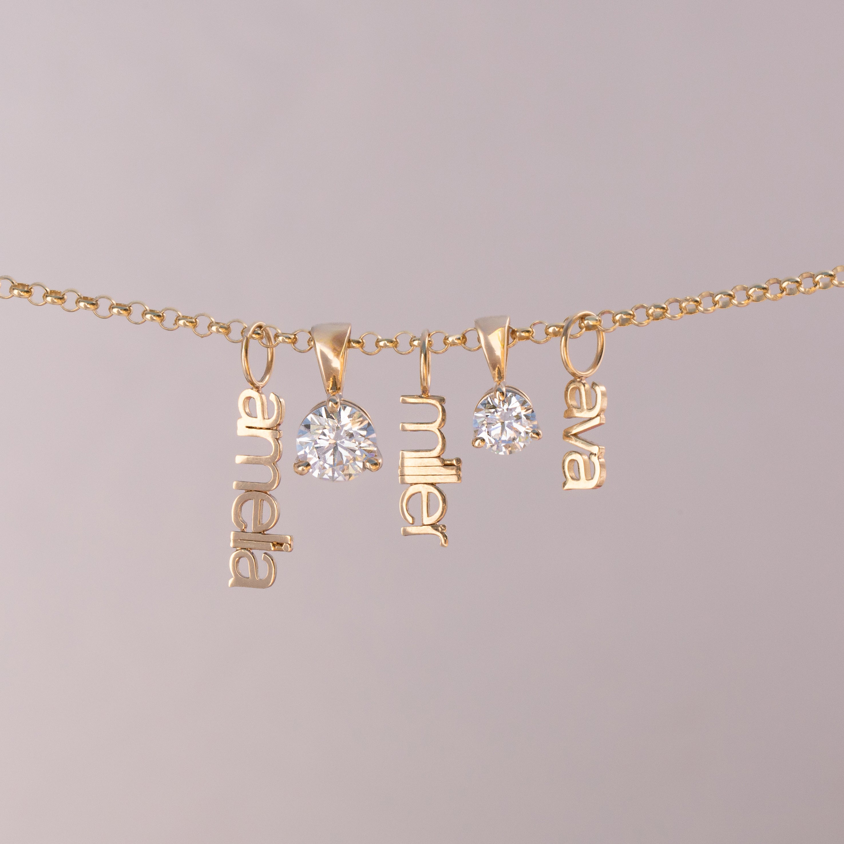 A gold chain necklace with three dangling nameplates spelling 'anabel', 'miller', and 'elva', alongside two clear round gemstones, shown against a solid light gray background.
