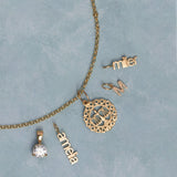 A gold jewelry set laid out on a textured light blue background, including a chain necklace with a circular scale of justice pendant, a solitaire diamond pendant, a nameplate reading 'anna', a nameplate reading 'miller', and a small nameplate reading 'M'.
