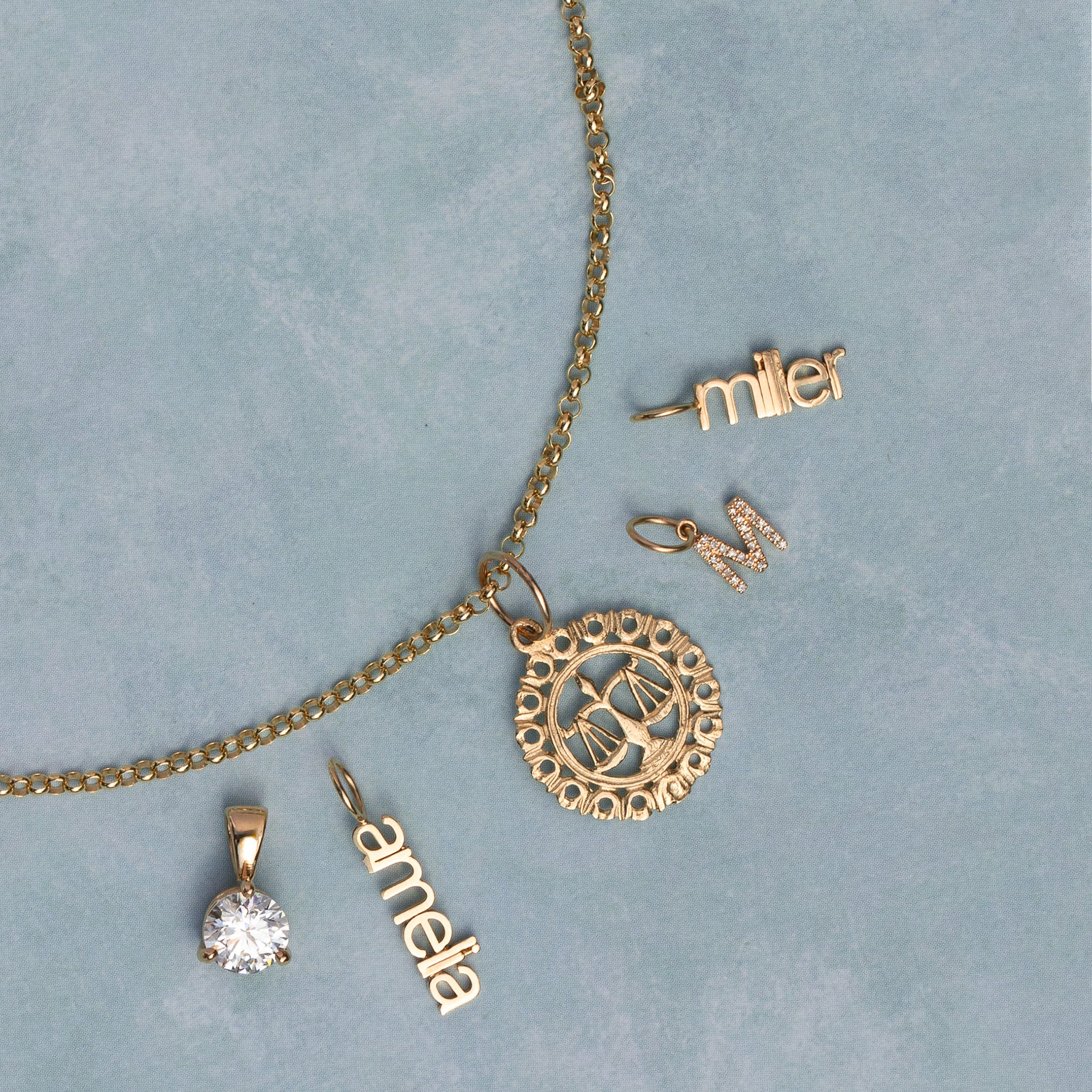 A gold jewelry set laid out on a textured light blue background, including a chain necklace with a circular scale of justice pendant, a solitaire diamond pendant, a nameplate reading 'anna', a nameplate reading 'miller', and a small nameplate reading 'M'.