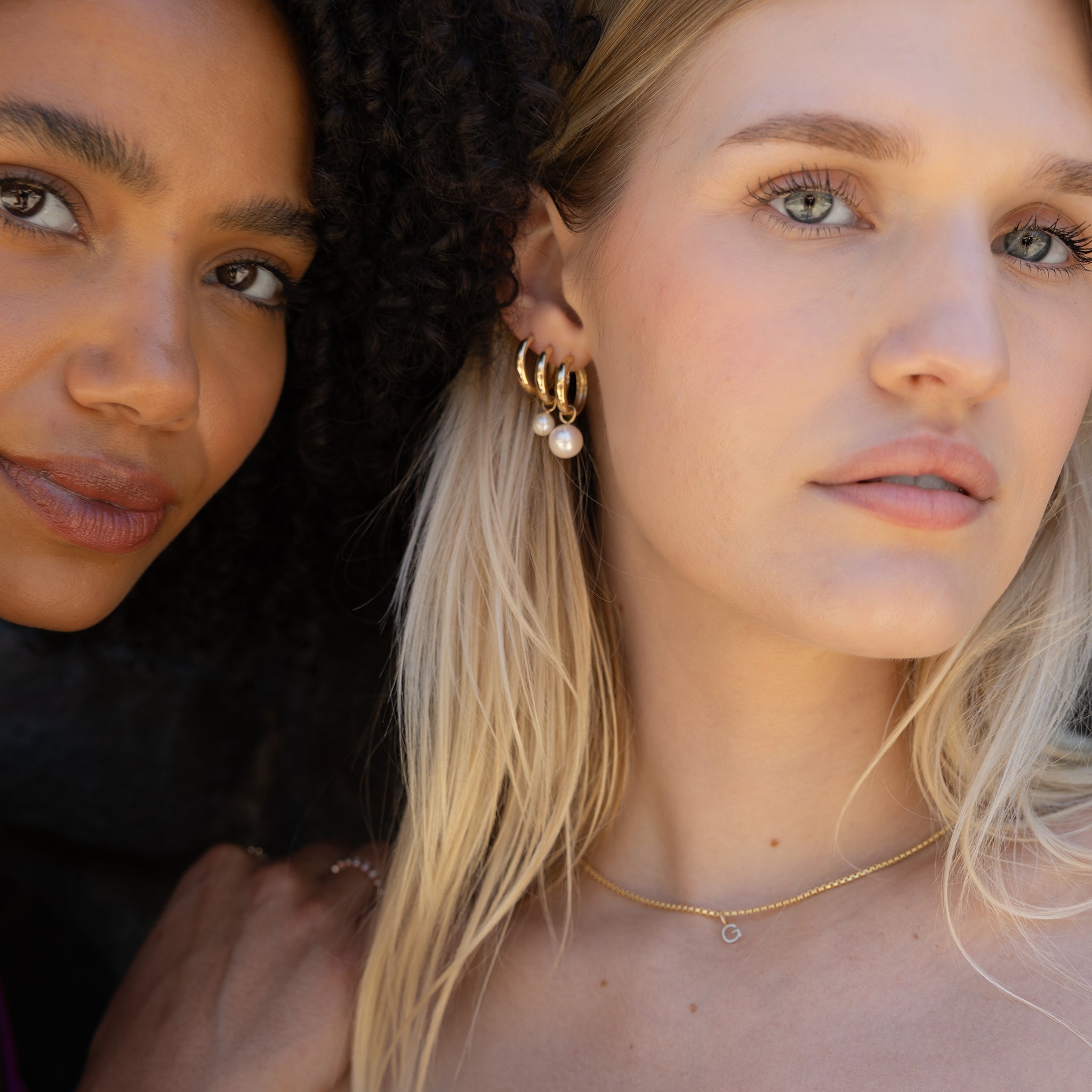 Close-up lifestyle shot of two women wearing gold jewelry, featuring a pair of gold hoop earrings with dangling white pearls and a delicate gold chain necklace.