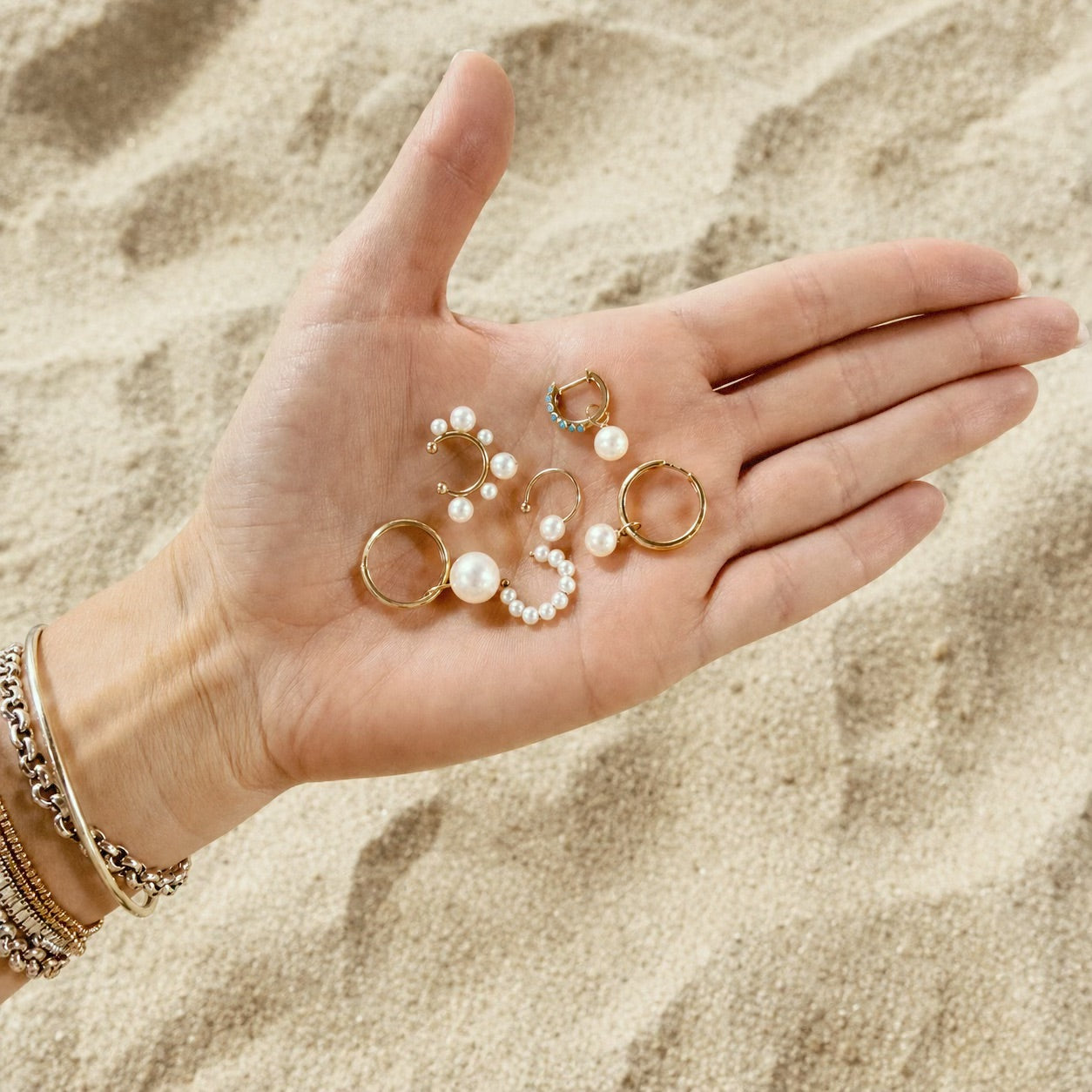 Assortment of Maya Brenner pearl hoop earrings and Soleil Hoops with pearl earring charms held in an open hand over sandy beach