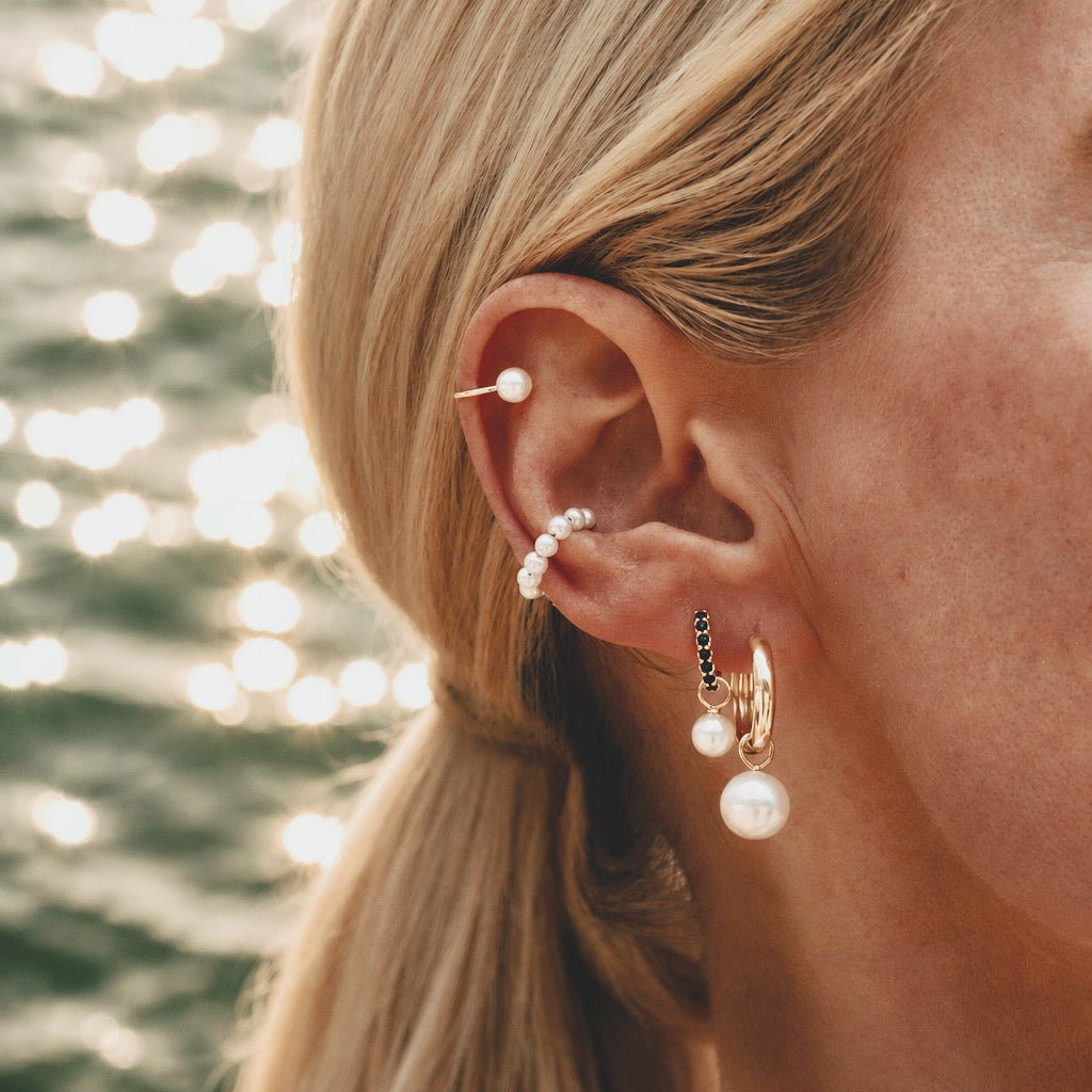 Model wearing Maya Brenner Solitaire Pearl Ear Cuff styled in a layered ear stack with pearl halo ear cuff, black diamond huggie hoops, and pearl earring charms