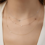 A close-up of a woman’s neck wearing  several Custom Gold Necklaces stacked: a two-letter white gold necklace with the letters C and M, a five-letter rose gold necklace with the letters D, L, H, and M and a birthstone, and a two-letter yellow gold necklace with the letters E and L.