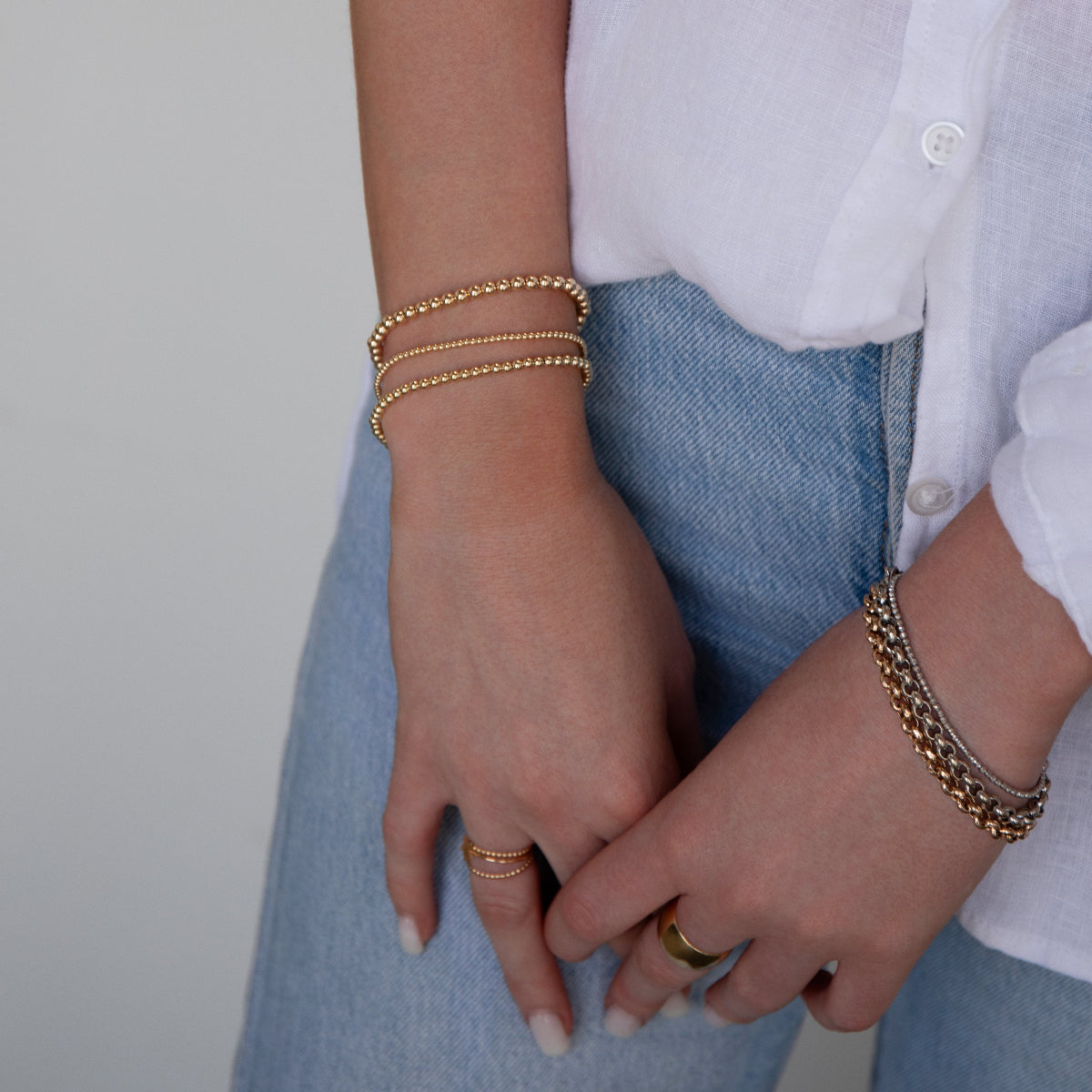 A close-up of a woman's hands wearing mixed metal jewellery, including a Classic Bubble Bracelet, Rolo Bracelets, a Diamond Cut Bracelet, Delicate Beaded Bands and a Thick Band, styled in a white shirt and light blue denim jeans. 