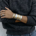 A close-up of an arm wearing several gold and silver All In Cuffs, Plus One Cuffs and Flirt Bangles, a white gold Etched Paris Ring and a Tres Pearl Ring, styled in a dark grey knit and blue jeans.