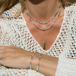 A close-up of a person wearing two Endless Summer Necklaces in Capri, The In Flow Retreat Necklace and two Endless Summer Bracelets with a cream crochet knit. 