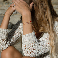 Close-up of woman's arm wearing two Endless Summer Bracelets in Capri and a Perla Pinky Ring, styled in a cream textured knit against a blurred stone background. 