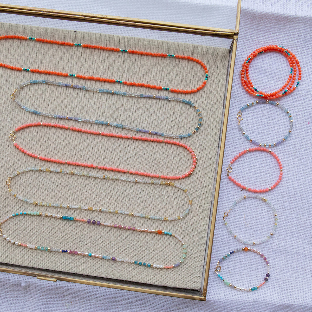 Endless Summer Necklaces and Bracelets (Calypso, Blue Lagoon, Mykonos, Saint-Tropez and Capri), with the necklaces laid in a clear tray on beige fabric and the bracelets laid next to the tray on a white fabric background.