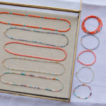 Endless Summer Necklaces and Bracelets (Calypso, Blue Lagoon, Mykonos, Saint-Tropez and Capri), with the necklaces laid in a clear tray on beige fabric and the bracelets laid next to the tray on a white fabric background.