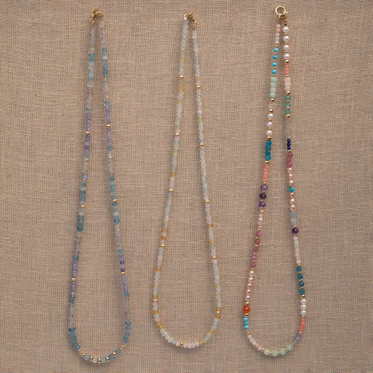 Three Endless Summer Necklaces (Blue Lagoon, Saint-Tropez and Capri) on a beige fabric background. 