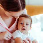 A woman wearing a MUM Necklace and a Geo Stacking Ring Square in yellow gold, holding a baby in a bedroom setting.  