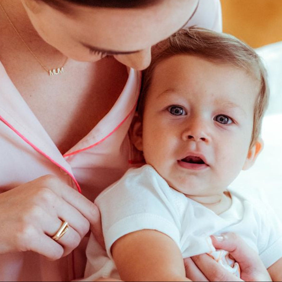 A woman wearing a MUM Necklace and a Geo Stacking Ring Square in yellow gold, holding a baby in a bedroom setting.  