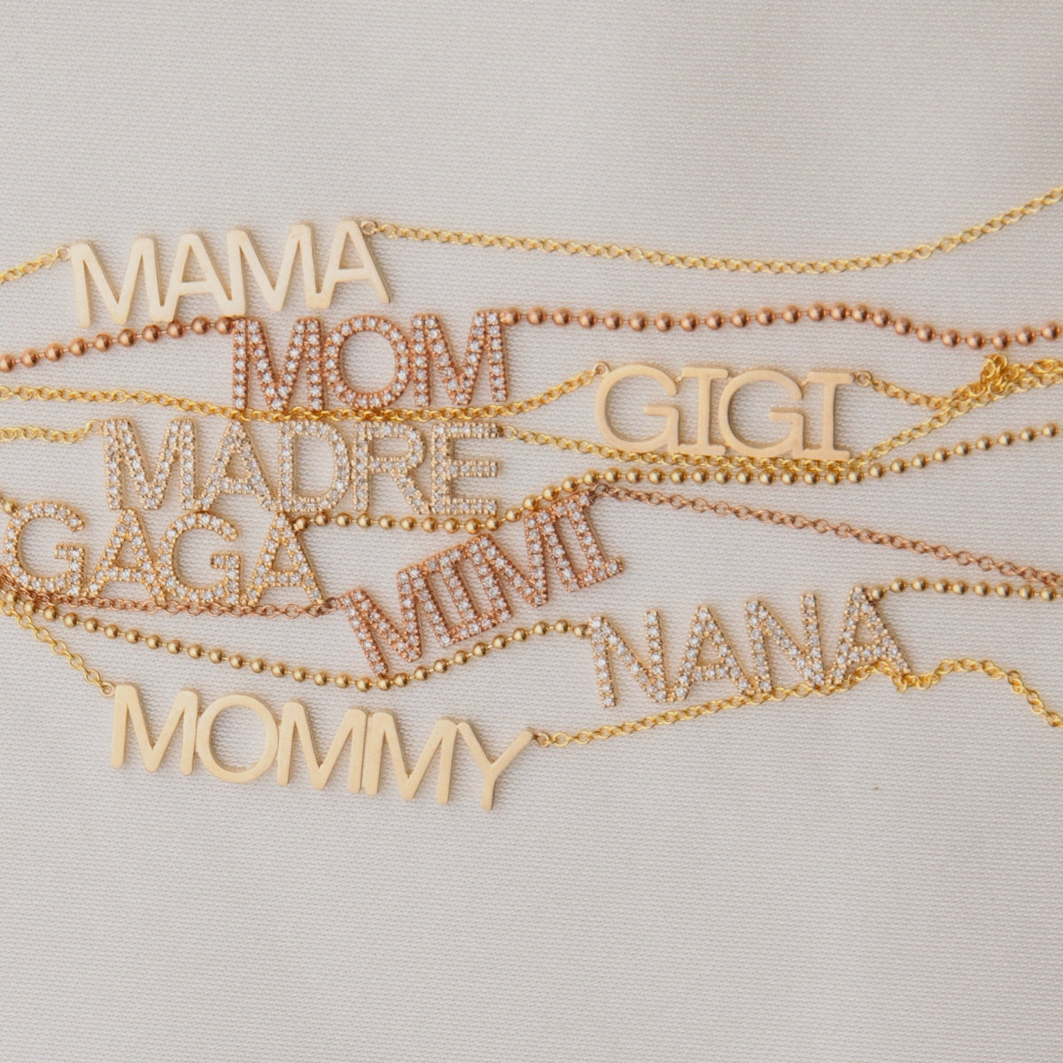 A MAMA Necklace, GIGI Necklace, Pavé MADRE Necklace, Pavé GAGA Necklace, Pavé NANA Necklace, and a MOMMY Necklace in yellow gold, along with Pavé MOM and MIMI Necklaces in rose gold, laid on an off-white background.