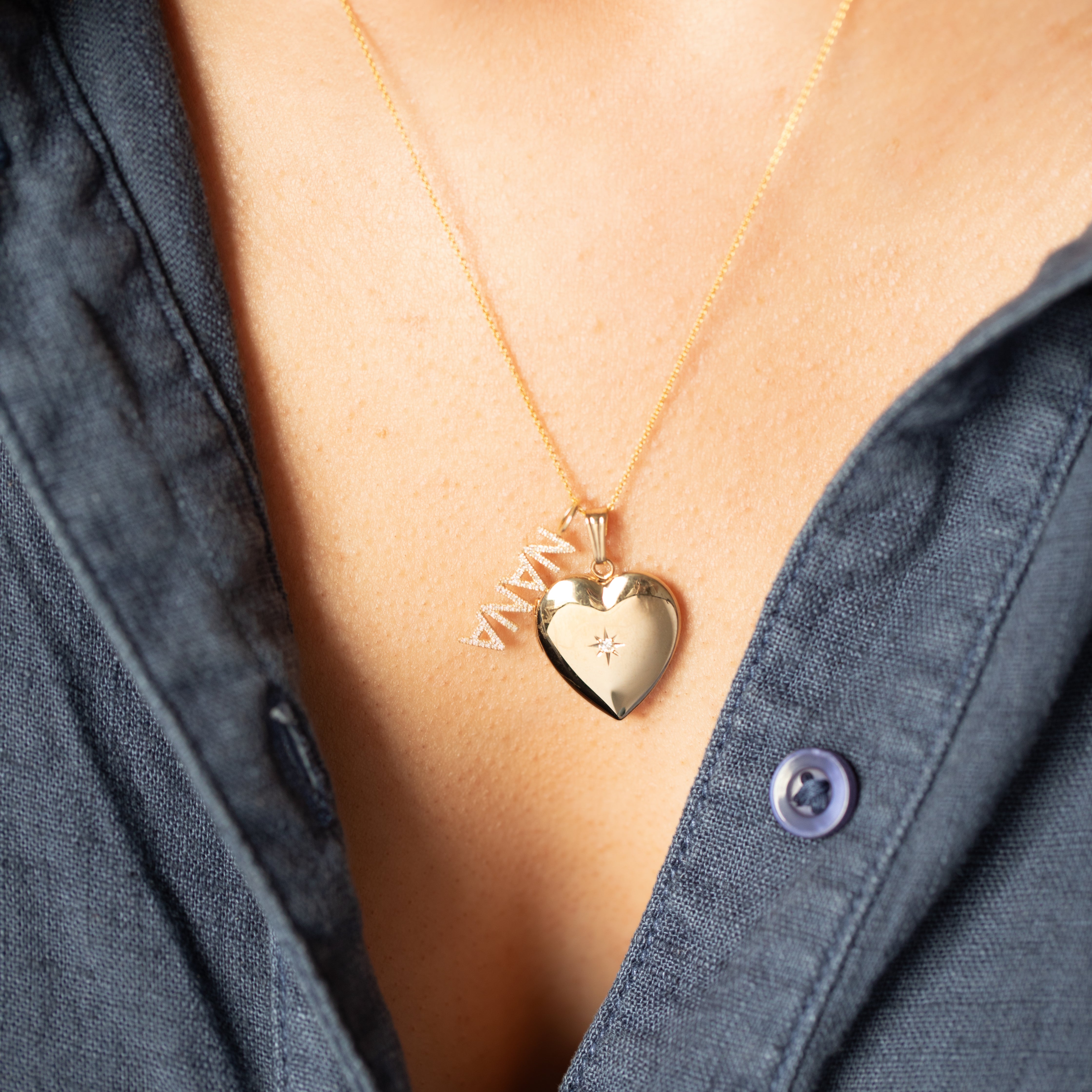 Gold heart-shaped necklace with diamond and pavé diamond 'NANA' pendant on a person wearing a blue shirt.
