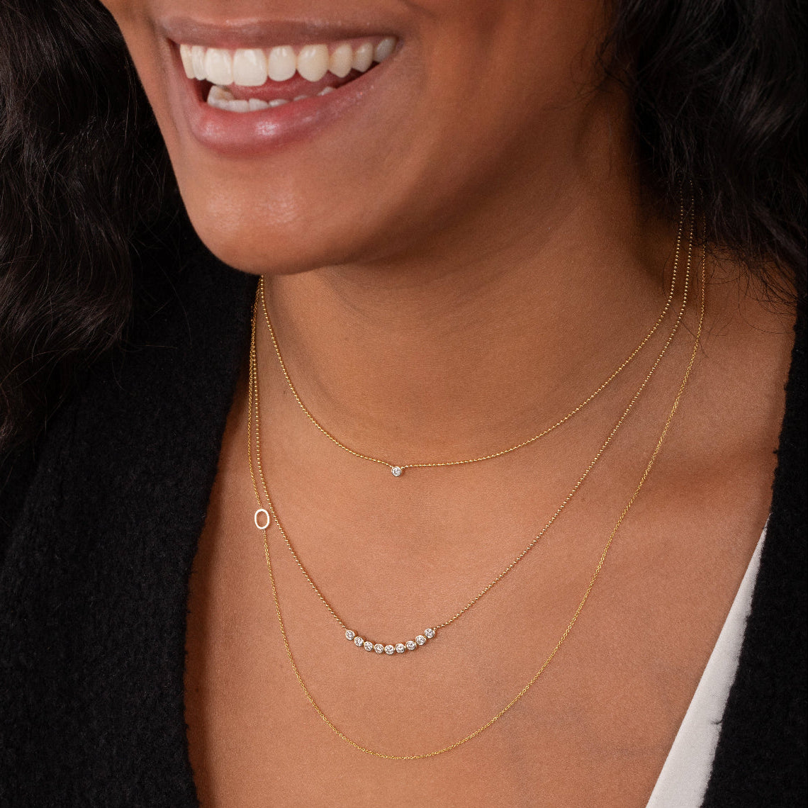 Close-up of a woman's neck wearing a Single Diamond Layering Necklace, a Diamond Arc Layering Necklace and a Custom Gold Necklace - 1 Letter, all in yellow gold, in a black top.