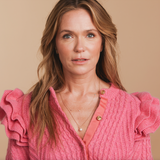 A woman wearing a Custom Gold Necklace with three birthstone charms and a Perla Necklace, styled in a pink sweater with ruffled sleeves against a beige background. 