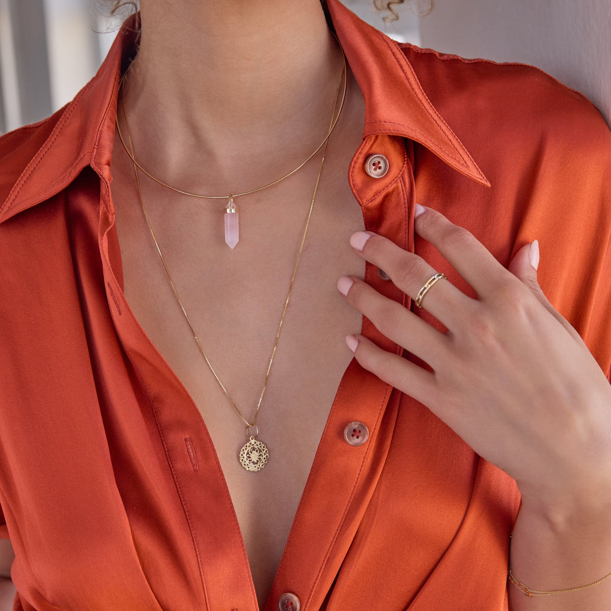 A woman's décolletage wearing an Omega Chain with a Balance Crystal Charm, Zodiac Necklace - Cancer and a Link Ring, with an orange silk shirt against a light neutral background. 