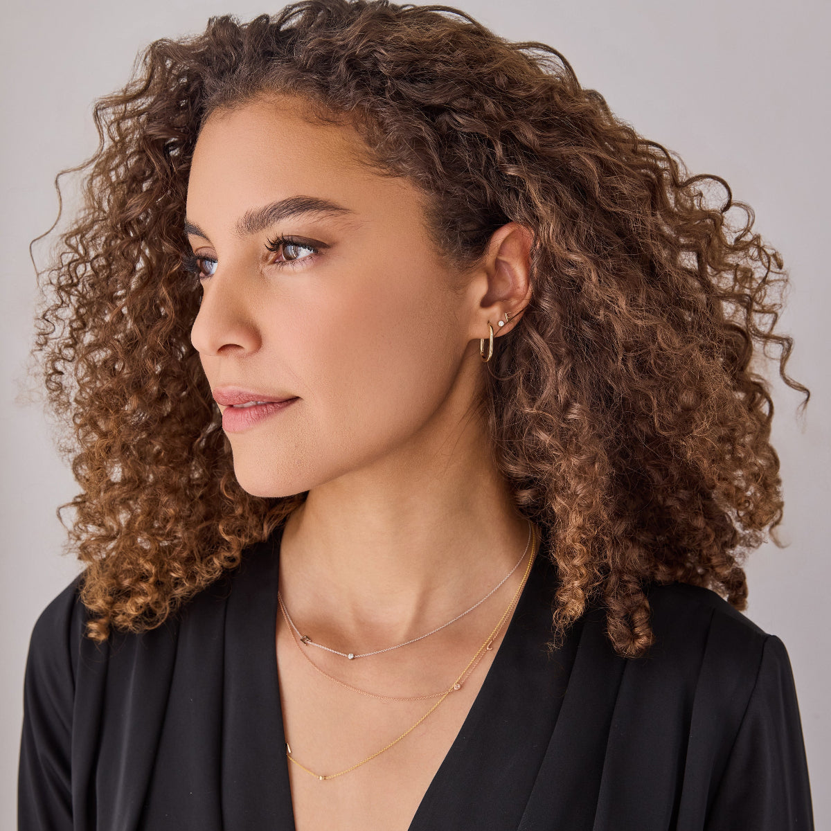 A woman wearing a yellow gold Oval Hoop, Monogram Earring Set and three Monogram Necklaces with Diamond, with a black top and brown curly hair, against a light grey background. 