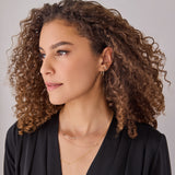A woman wearing a yellow gold Oval Hoop, Monogram Earring Set and three Monogram Necklaces with Diamond, with a black top and brown curly hair, against a light grey background. 