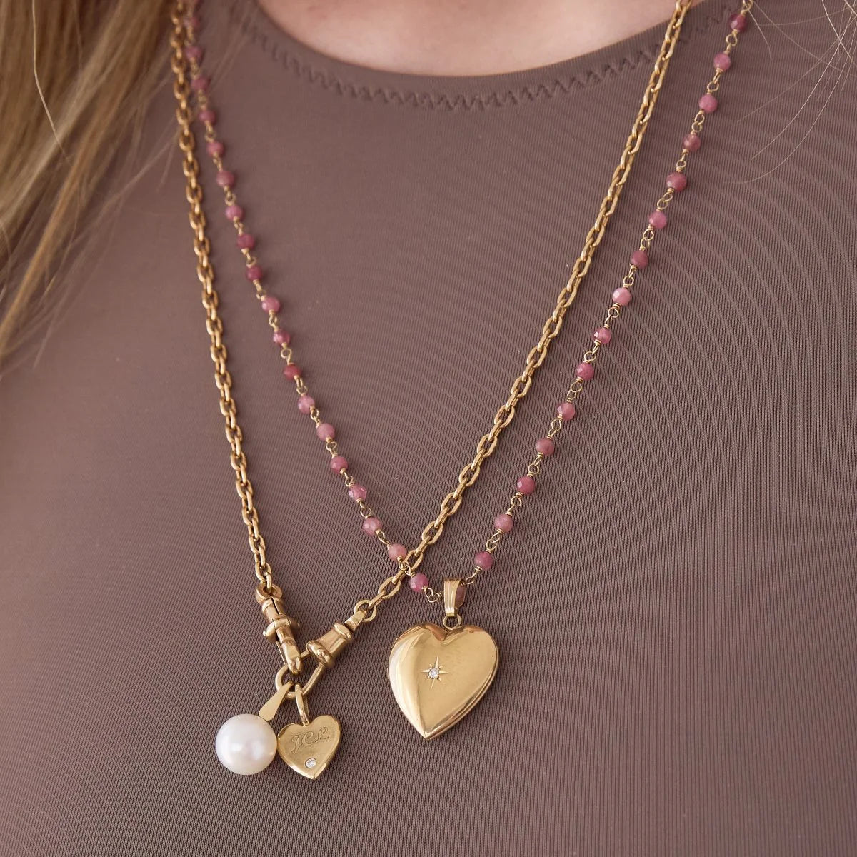 Close-up of a person's decellotage wearing an Oval Link Chain with a Perla Pendant and Engraved Heart Charm and  The Peony Bouquet Necklace with a Diamond Heart Locket Charm, styled in a brown top. 
