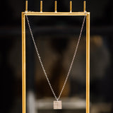 A Virtues Pendant - Square in sterling silver hung on a gold jewellery stand against a dark blurred background. 