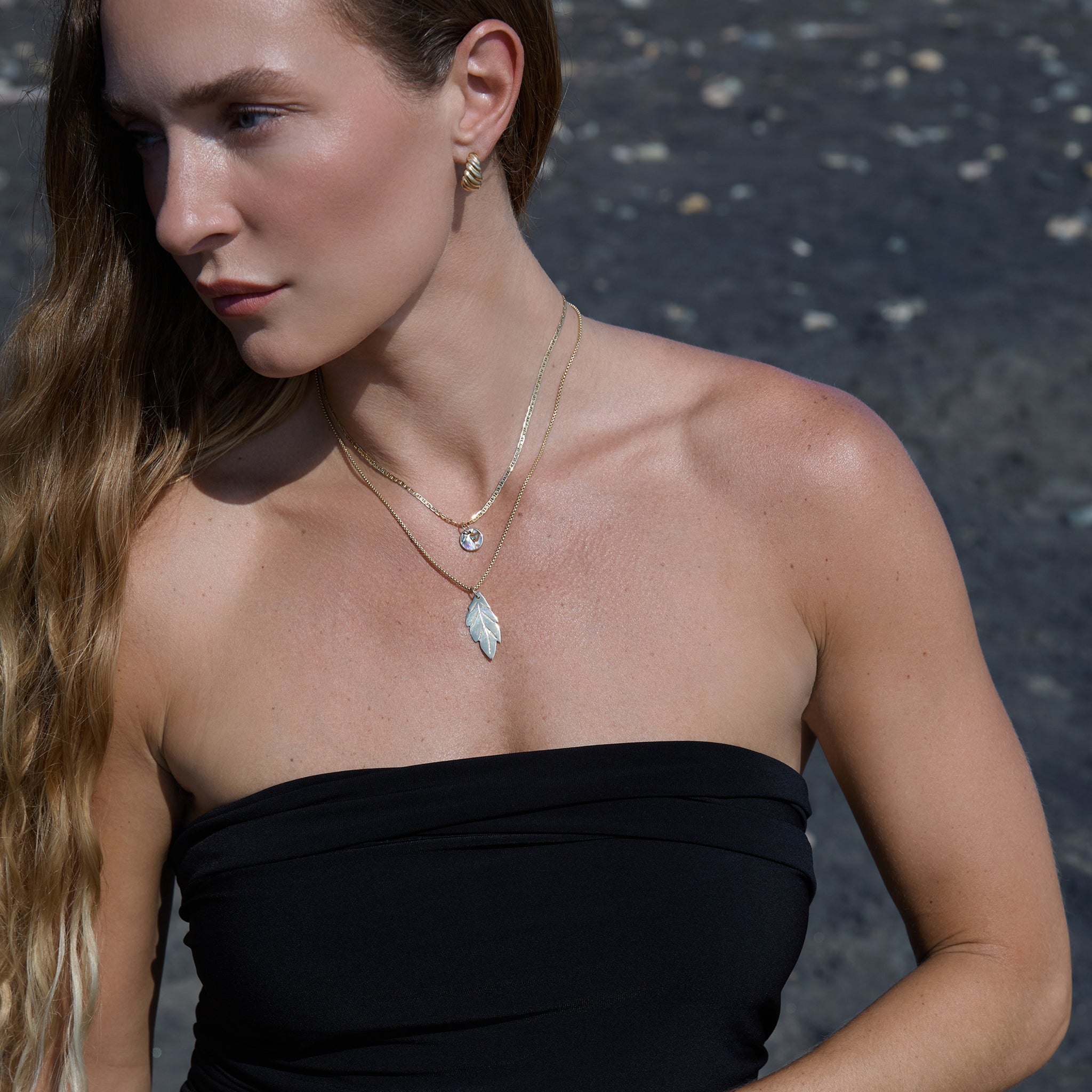 Woman wearing a black strapless top with layered gold charm necklaces against a dark background