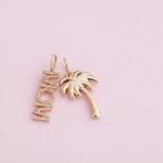 A pair of gold-toned jewelry charms on a soft pink background. One charm is a vertical script word 'MOM' set with small clear stones, and the other is a palm tree silhouette in polished gold.