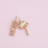A pair of gold-toned jewelry charms on a soft pink background. One charm is a vertical script word 'MOM' set with small clear stones, and the other is a palm tree silhouette in polished gold.
