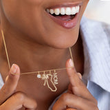 A close-up lifestyle shot of a woman holding a delicate gold necklace with charms. The necklace features a small clear gemstone, a palm tree, a California state silhouette, and the word 'MOM'.