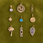 Nine gold-toned jewelry pendants arranged in a 3x3 grid on a textured olive green background. The pendants include a round clear gemstone, a circular filigree pendant with a lion motif, a script nameplate reading 'ava', a small flower with clear stones, a round blue gemstone pendant, a circular medallion with a sun motif, a palm tree pendant, a vertical 'MOM' nameplate with clear stones, and a leaf-shaped pendant with a purple and blue inlay.