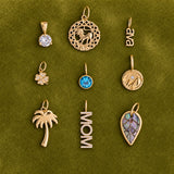 Nine gold-toned jewelry pendants arranged in a 3x3 grid on a textured olive green background. The pendants include a round clear gemstone, a circular filigree pendant with a lion motif, a script nameplate reading 'ava', a small flower with clear stones, a round blue gemstone pendant, a circular medallion with a sun motif, a palm tree pendant, a vertical 'MOM' nameplate with clear stones, and a leaf-shaped pendant with a purple and blue inlay.