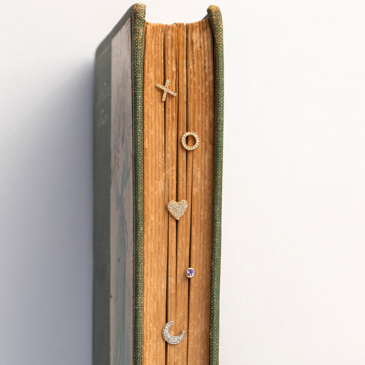 Pavé Charm Studs - Heart and Moon, a Birthstone Stud and two 14K Gold Pave Letter Studs in X and O displayed on top of a book against a white background. 