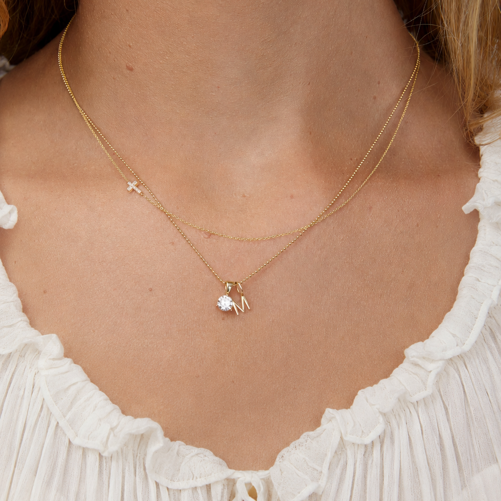 A woman's neck wearing a yellow gold Classic Bead Chain with a Gold Letter Jumble - M, and a 14k Gold Asymmetrical Charm Necklace - Pave Cross.