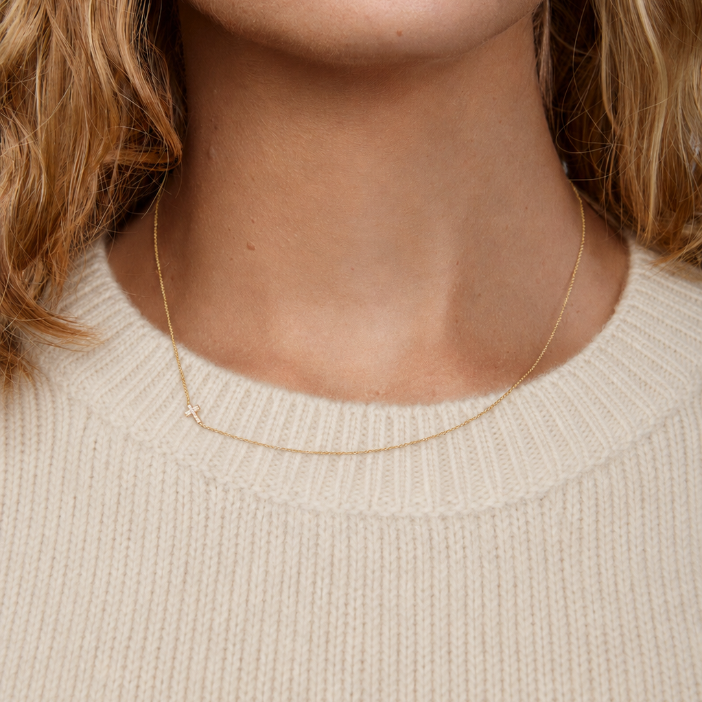 Close-up of a person wearing a delicate gold Cross necklace against a cream sweater.