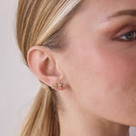 Close-up of Pavé Charm Studs - Cross and Om in yellow gold in a blonde woman's ear against a light grey background. 