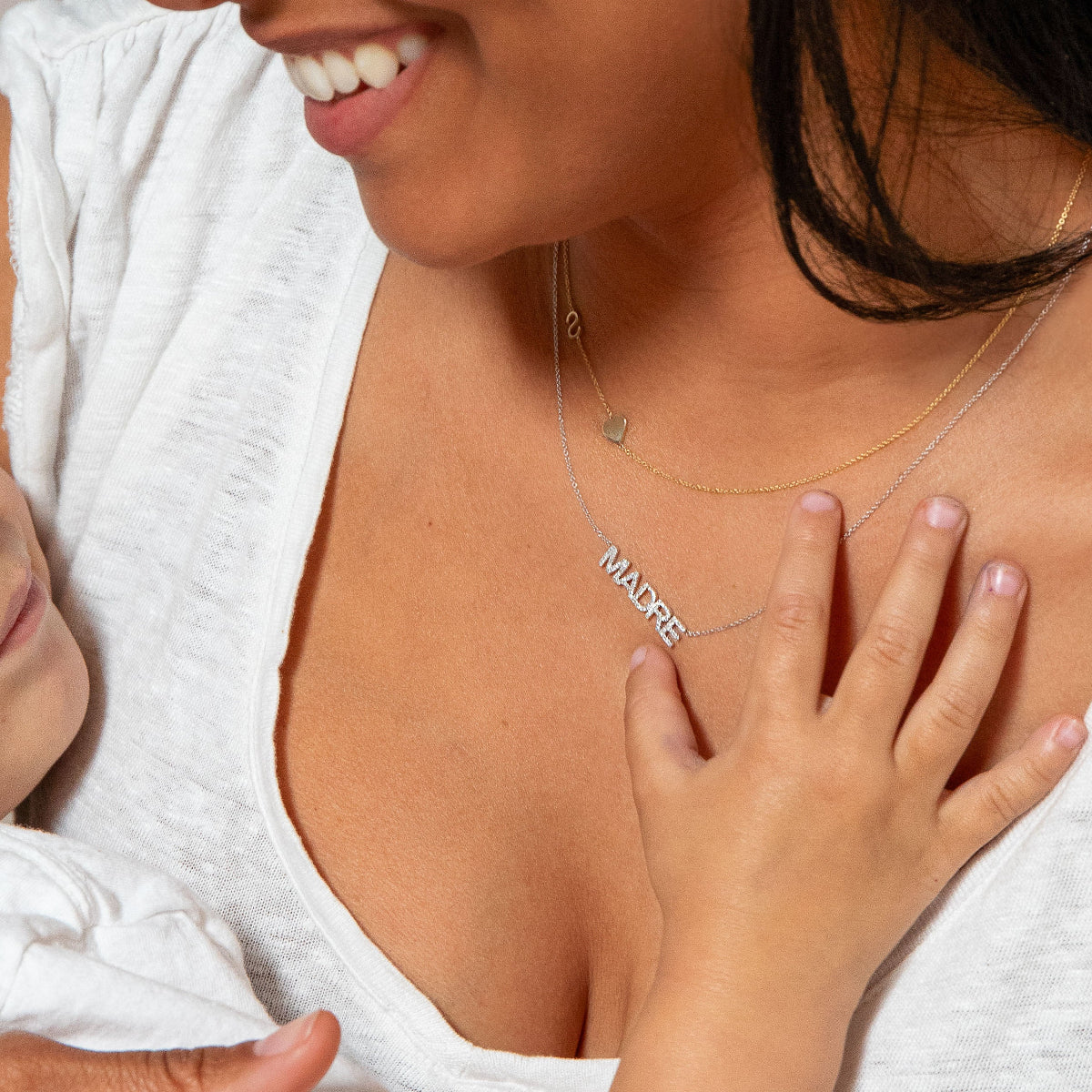 Close-up of a woman's neck wearing a yellow gold Custom Gold Necklace — 2 Letters (S and a heart charm) and a white gold Pavé MADRE Necklace, in a white T-shirt, smiling and holding a child.