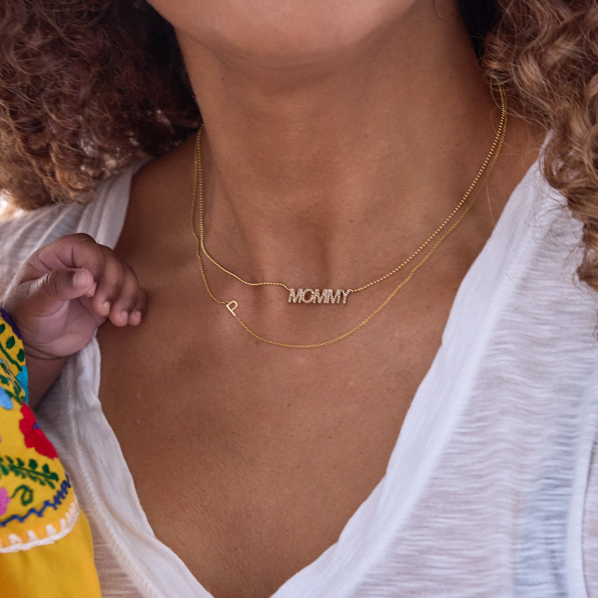 Close-up of a woman's neck wearing a Pavé MOMMY Necklace and a Custom Gold Necklace - 1 Letter (P), both in yellow gold, in a white T-shirt with brown curly hair visible, with a child's hand resting on her shoulder.
