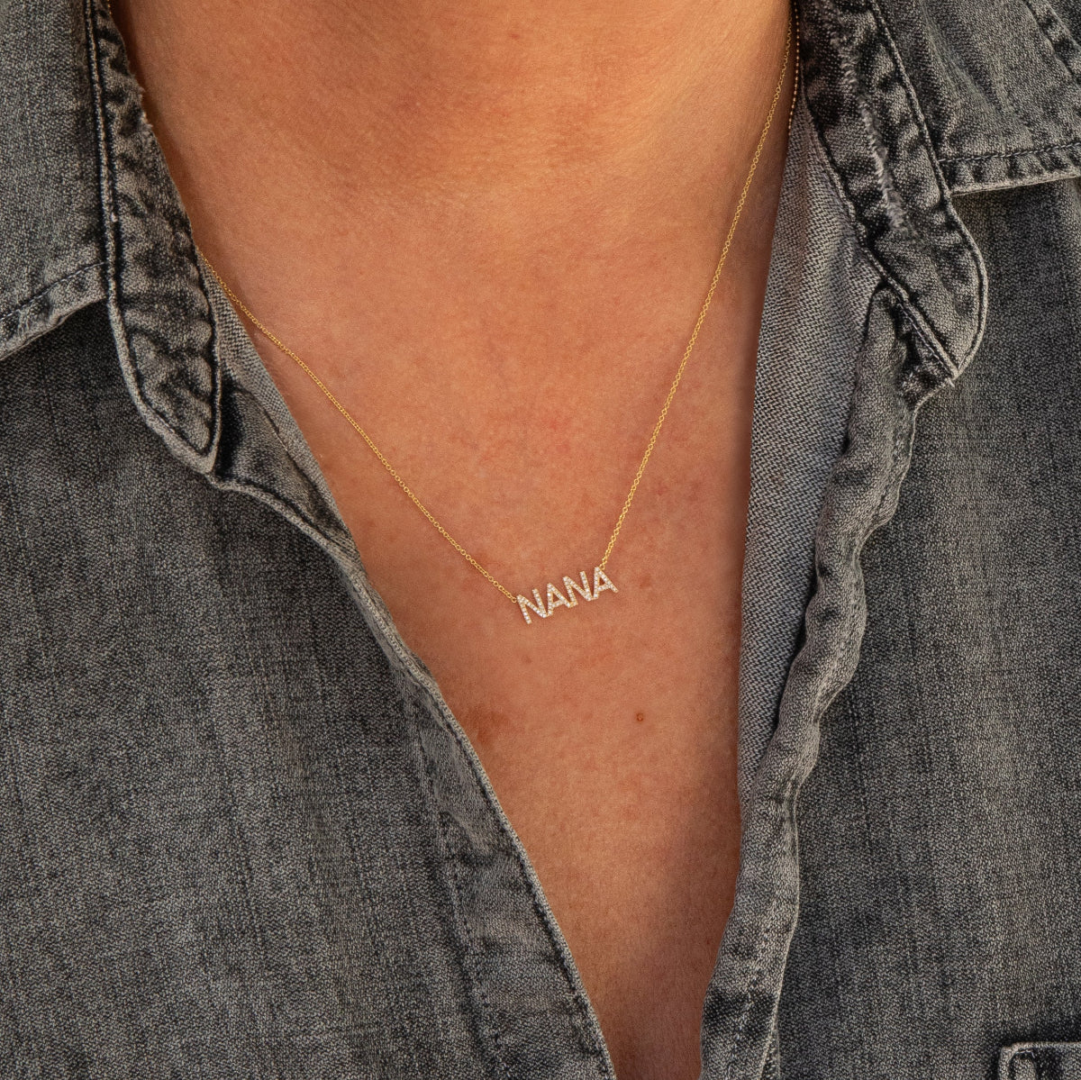 A close-up of a décolletage wearing a Pavé NANA Necklace in yellow gold and a cable chain, with a grey denim shirt. 
