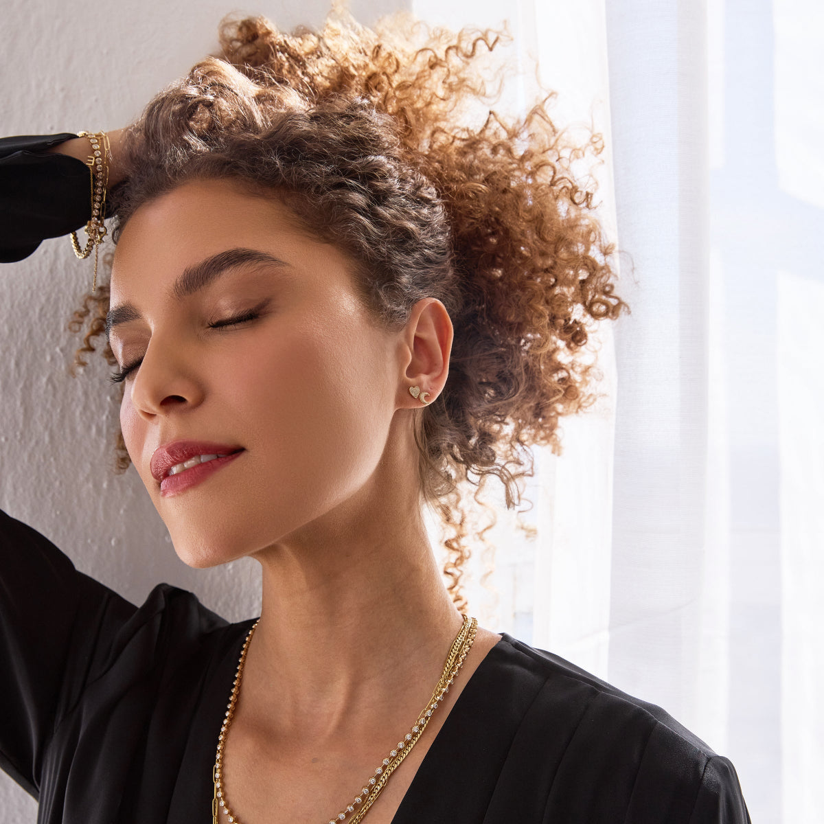 A woman wearing a Pavé Charm Stud - Heart and Moon, The Ultimate Diamond Necklace, Chic Cuban Curb Chain, Love Set Match Bracelet, Element Long Link Bracelet, and Custom Gold Bracelet - 1 Letter, all in yellow gold, with brown curly hair and a black top against a white background.