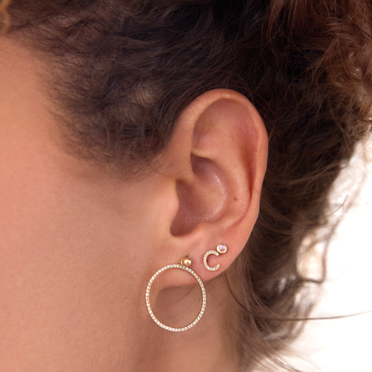 Close-up of an ear wearing a Birthstone Stud - Moonstone (June), 14K Gold Pavé Letter Stud in C and a Mini Preppy Post with a Circle Ear Jacket Pave, against a white blurred background. 