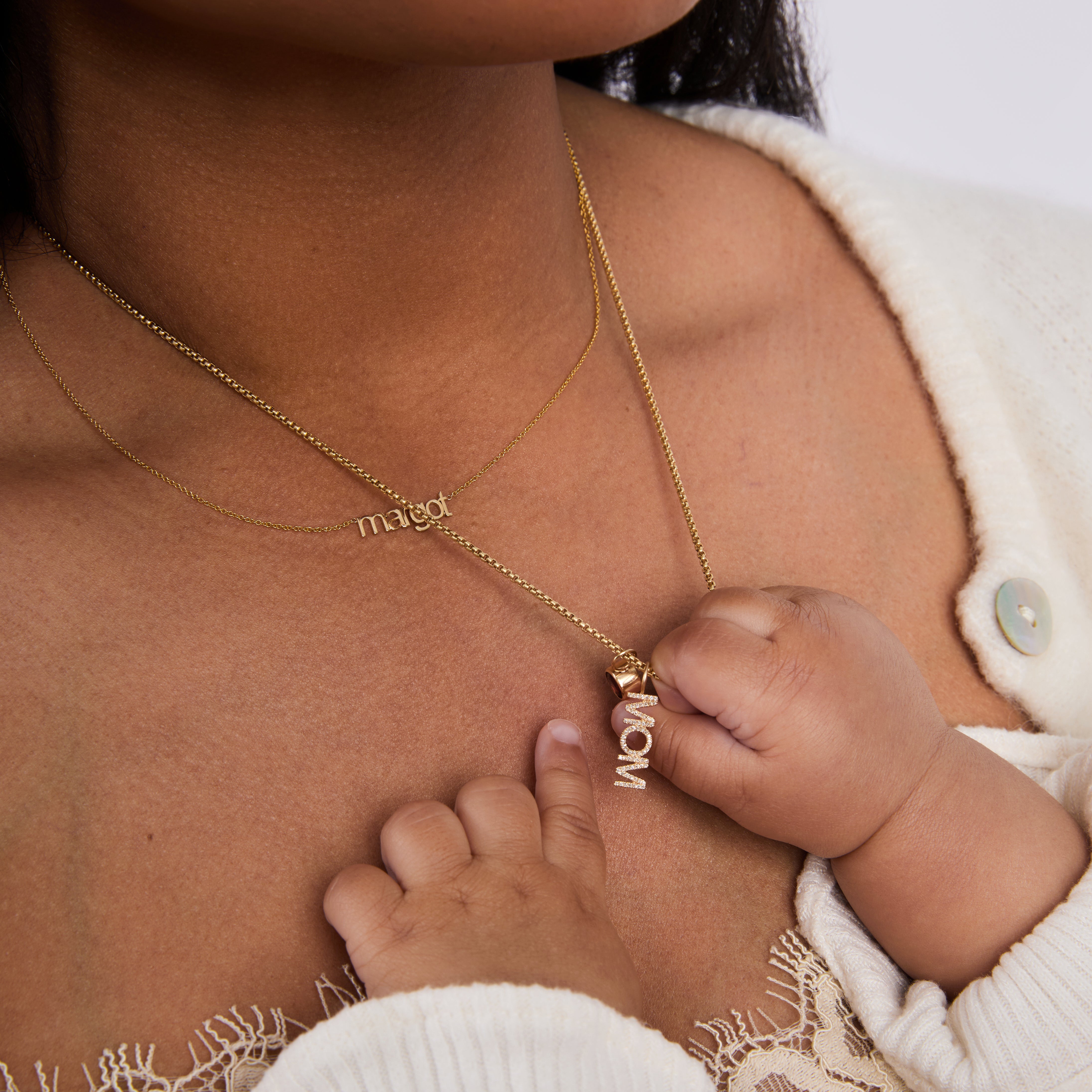 Close-up of a person wearing a gold necklace with a baby's hand holding a pavé 'mom' pendant.