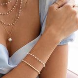A close-up of a woman’s arm and décolletage wearing three layered The Lily Bouquet Necklaces in different lengths, a Perla Necklace, The Lily Bouquet Bracelet and a Little Bubble Bracelet, styled with a light blue top.