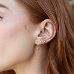 A close-up of a woman's ear wearing two Birthstone Studs (Diamond and Pearl) and a Gold Safety Pin Earring, with orange hair. 