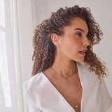 A woman wearing a Tres Pearl Earring, Bold Link Chain, MOM Necklace and Perla Necklace, with brown curly hair and a white top against a white background. 