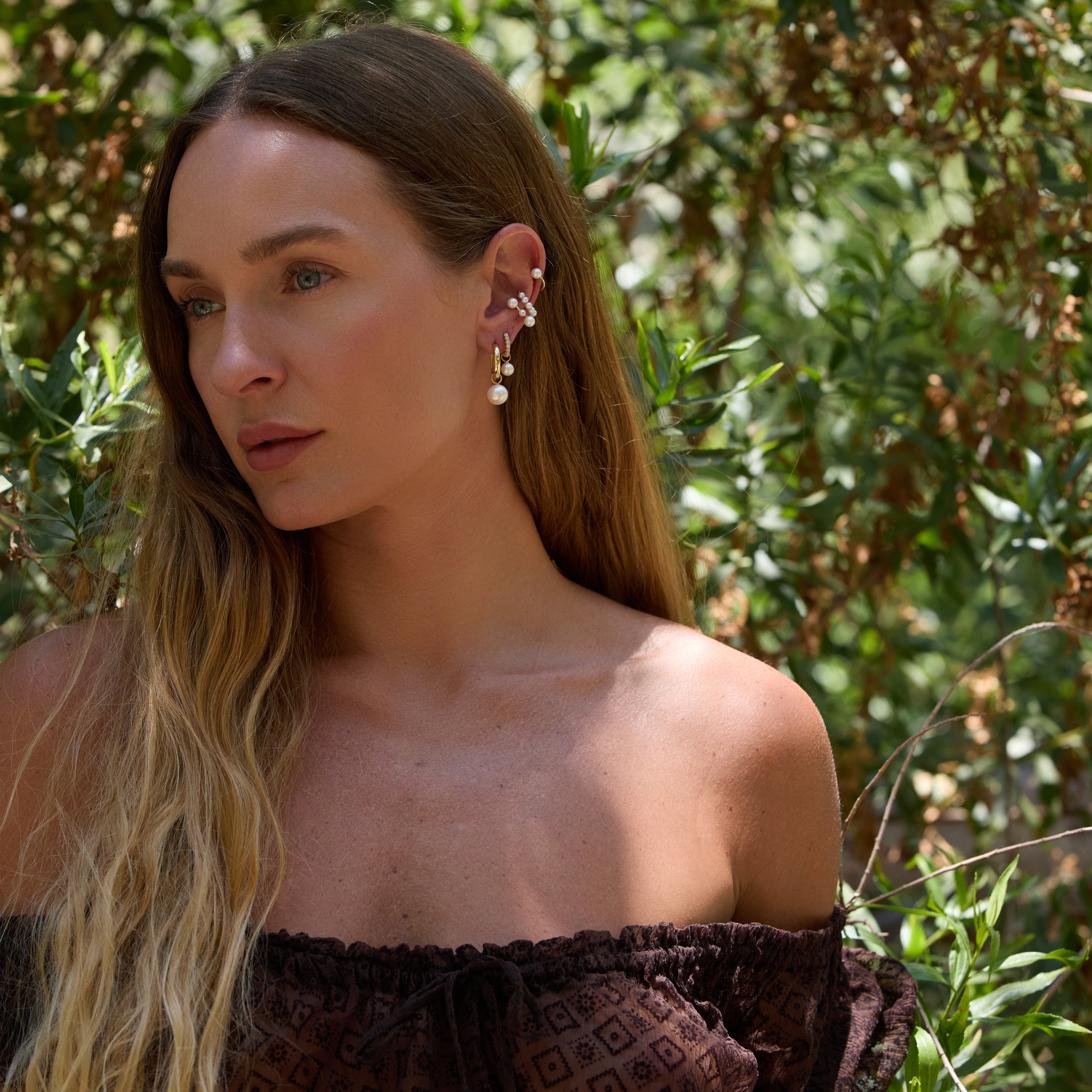Woman with long hair and pearl earrings standing outdoors with greenery in the background