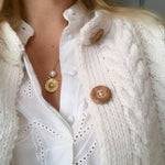 Close-up of a blonde woman's décolletage, wearing a Perla Necklace paired with another gold necklace, styled in a white shirt and cable knit cardigan with wooden buttons. 