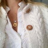 Close-up of a blonde woman's décolletage, wearing a Perla Necklace paired with another gold necklace, styled in a white shirt and cable knit cardigan with wooden buttons. 