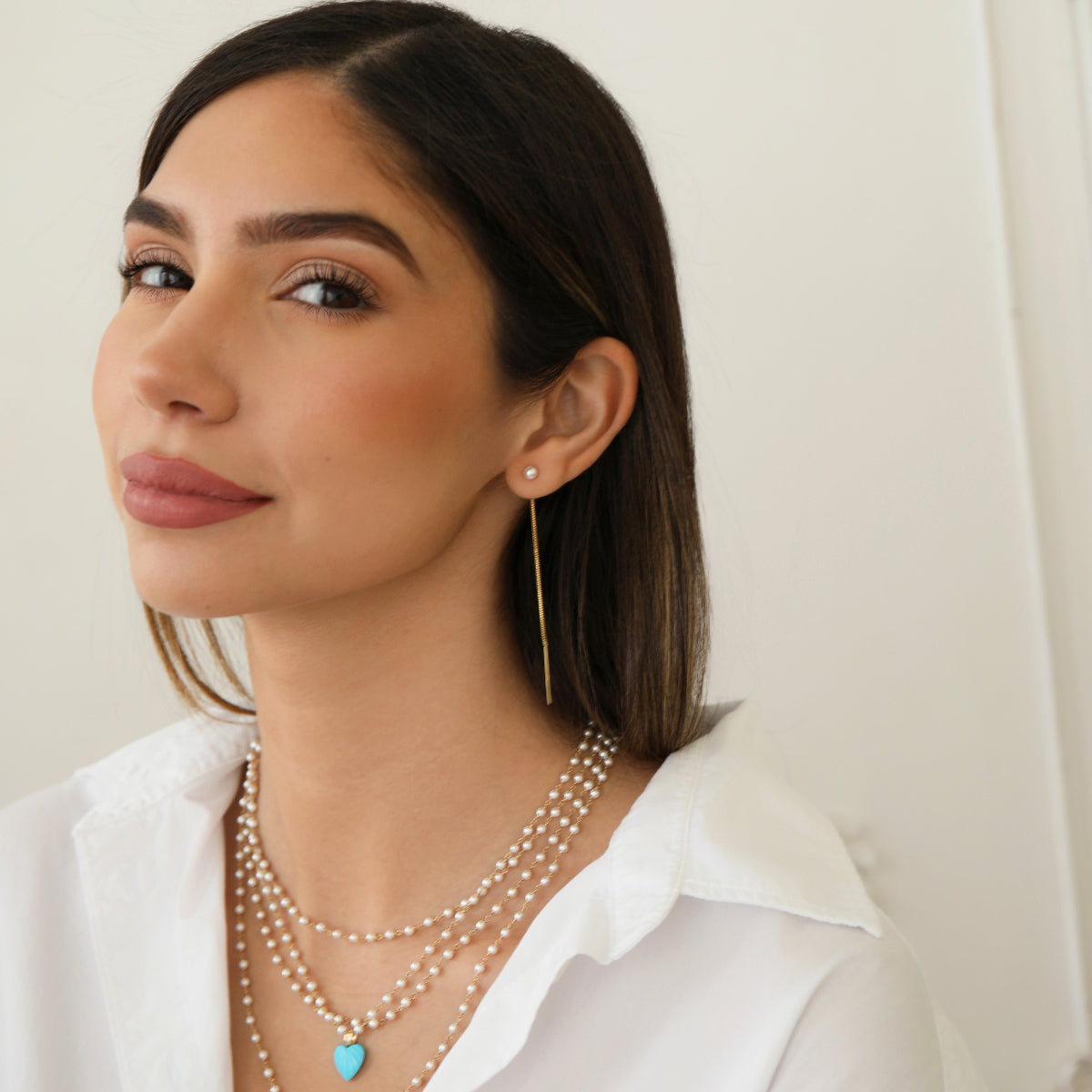 A woman wearing a Pearl Drift Threader, several The Lily Bouquet Necklaces and a white top, with brown hair against a neutral background.