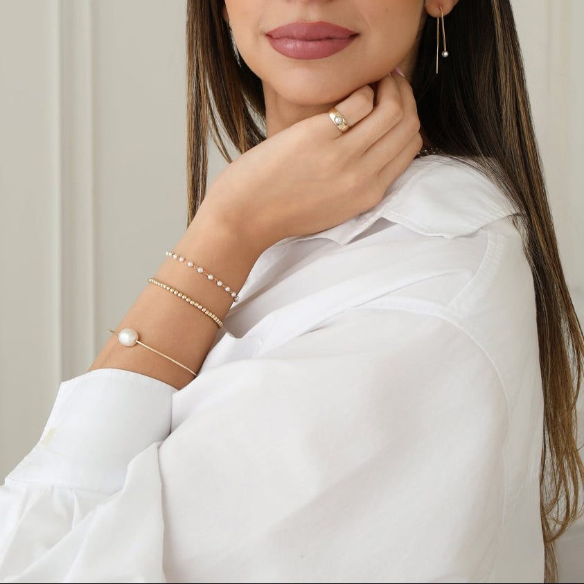 A woman wearing The Lily Bouquet Bracelet, Baroque White Pearl Bangle, Little Bubble Bracelet, Perla Pinky Ring and Pearl Drift Threader, styled with a white shirt. 
