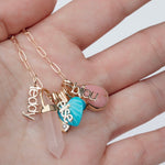 A hand holding an Element Short Link Chain with several charms, including Petite Nameplate Pendants 'bijou', 'teddy' and 'elise', an Etched Turquoise Heart Charm, a Balance Crystal Charm, an Engraved Heart Charm and an Engraved Heart Charm. 